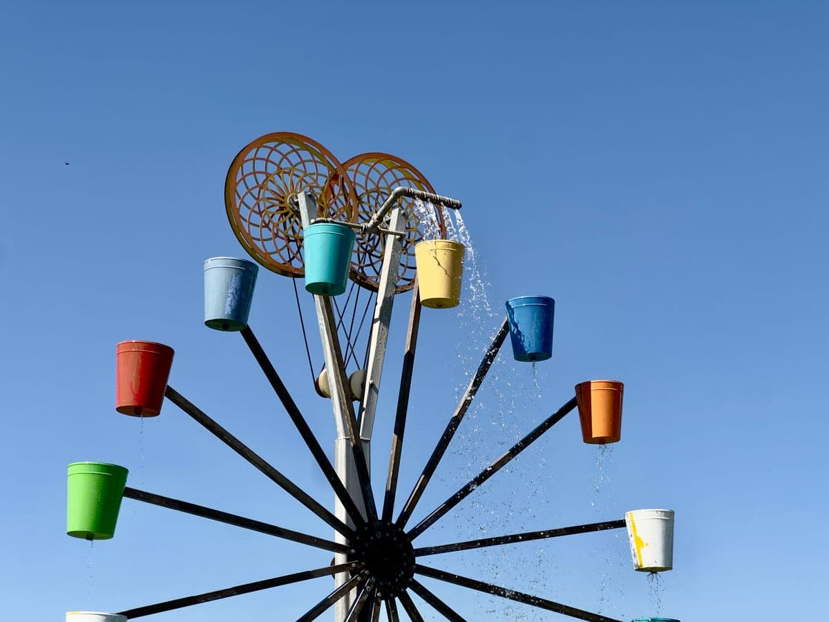 A water-wheel sculpture with colored buckets on its spokes, turned by water poured from a pipe